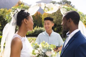 Happy diverse male officiant with bride and groom at outdoor wedding ceremony in sunny garden